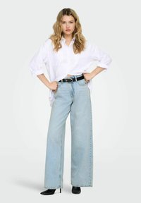 Chemise blanche boutonnée, jean bleu clair à jambes larges, ceinture noire avec boucle circulaire et bottines noires à talons. Tissu lisse et coupe ample.