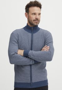 Suéter de punto en azul con cuello alto y cremallera frontal. Presenta rayas verticales y puños y dobladillo acanalados para mayor textura.