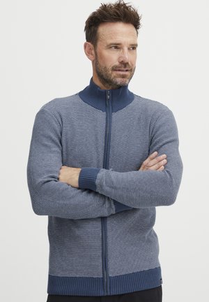 Strikket genser i blått med høy krage og glidelås foran. Har vertikale striper og ribbestrikkede mansjetter og kant for ekstra tekstur.