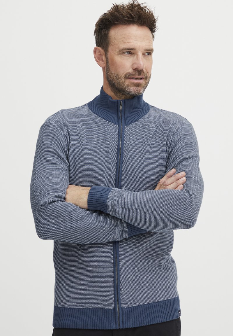 Suéter de punto en azul con cuello alto y cremallera frontal. Presenta rayas verticales y puños y dobladillo acanalados para mayor textura.
