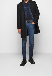 Cappotto scuro su una giacca di denim, polo blu navy e jeans blu; scarpe nere; look casual a strati con diverse trame di tessuto.