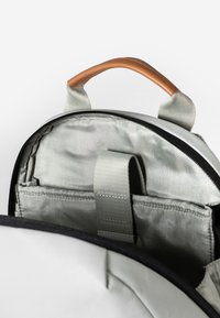 Rucksack mit einer glatten, hellgrauen Außenseite, kontrastierenden schwarzen Reißverschlüssen, einem handlichen, braunen Ledergriff und mehreren inneren Stauraumfächern mit einem metallischen Glanz.