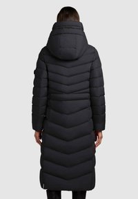 Lång svart pufferjacka med ett zigzag-mönster, huva och långa ärmar. Materialet ser mjukt och vadderat ut, med sidofickor synliga.