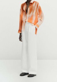 Chemise à manches longues avec un motif de coups de pinceau orange et blanc, associée à un pantalon large blanc et des sandales noires. Texture de tissu lisse.