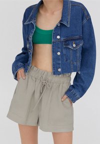 Veste en jean à coupe courte, couleur bleu clair, boutons argentés, associée à un top court côtelé vert et un short beige à cordon avec poches.