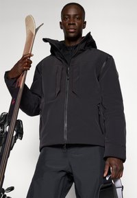 Schwarze isolierte Skijacke mit zwei Fronttaschen, einem hohen Kragen und einer Kapuze. Das Modell hält Skier und trägt schwarze Hosen. Glatte Textur.