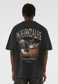 Fekete, oversize póló, élénk sassal és villámokkal körülvéve, a "M. GONZALES" felirattal felette és egy idézettel alatta.