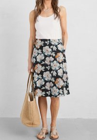 Woman wearing a white tank top, black floral knee-length skirt, beige sandals, carrying a large woven beige tote bag.