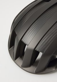 Cască de ciclism neagră cu un design elegant, aerodinamic, având fante de ventilație largi, textură mată și accente cu model din fibră de carbon.