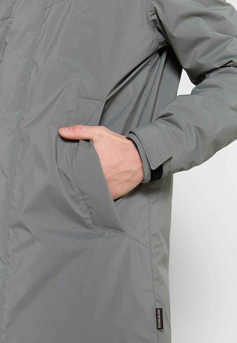 Giacca esterna grigia con texture liscia, chiusura con zip e tasche laterali. Include un piccolo'etichetta con il marchio vicino all'orlo.
