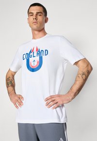 Hvid bomulds T-shirt med blå og røde "ENGLAND"-tekst samt et cirkulært grafik. Korte ærmer og klassisk crew neckline.