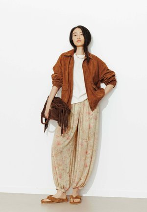 Jeune femme portant une veste en daim marron, un pantalon ample à fleurs, portant un sac marron à franges et des sandales marron, debout contre un mur blanc.