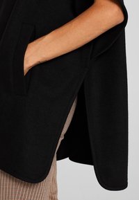 Ręka osoby w kieszeni czarnego płaszcza z tkaniny, nosząca wzorzyste brązowe spodnie, a płaszcz ma detal w postaci bocznego rozcięcia.