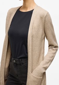 Beige knitted cardigan with open front, ribbed cuffs, and front pockets, worn over a black top. Features a soft texture and relaxed fit.