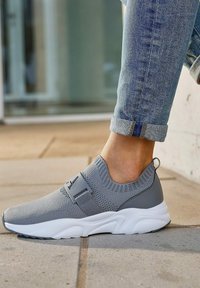 Grauer Sportschuh mit einem gestrickten Obermaterial, einem Akzentriemen und einer weißen Sohle. Glatte Textur, niedrig geschnittenes Design, ideal für die Freizeit.
