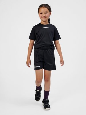 Młoda dziewczyna idzie do przodu, ubrana w czarną sportową odzież hummel, fioletowe skarpetki i czarne trampki na jednolitym jasnym tle.