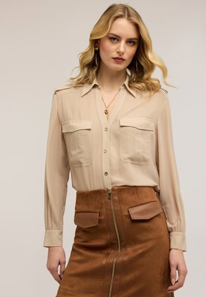 Vrouw met golvend blond haar, gekleed in een beige overhemd met knopen en zakken, en een bruine suède rok met een voorrits en klepzakken.