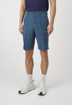 Mand iført blå knælange shorts, hvide sokker og hvide sneakers, står mod en ensfarvet lys baggrund.