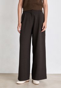 Pantaloni a gamba larga marrone scuro realizzati in tessuto testurizzato, con una piega frontale, vita alta e vestibilità su misura. Indossati con scarpe bianche.