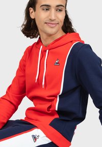Felpa con cappuccio rossa e blu navy con pannelli a contrasto, cappuccio con cordino e logo decorativo. Materiale morbido con vestibilità comoda e polsini a coste.