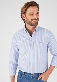 Camicia button-up a righe azzurre e bianche in cotone. Presenta un colletto classico e maniche arrotolate, abbinata a jeans in denim.