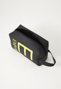 Bolsa de cosméticos preta com um fecho, apresentando uma marcação amarela em destaque de um lado e um acabamento texturizado. Inclui uma pega plana para transporte.