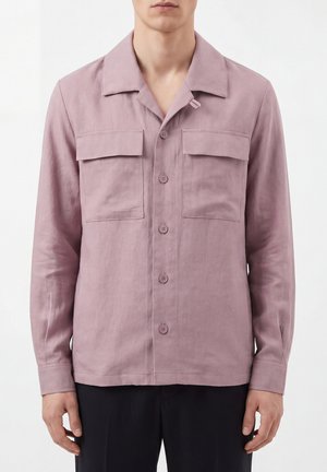 Man draagt een lange mouwen stoffig roze overhemd met twee borstzakken en een kraag, gecombineerd met een donkere broek.