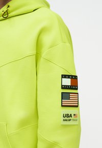 Φωτεινό κίτρινο-πράσινο μανίκι φούτερ με τρία μπάλωμα: λογότυπο Tommy Hilfiger, σημαία ΗΠΑ και κείμενο "USA SAIL GP TEAM".