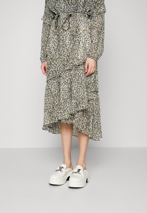 Person trägt ein midilanges Kleid mit Rüschen und Leopardenmuster sowie weiße Plateauslipper mit silbernen Schnallendetails und steht vor einem schlichten Hintergrund.
