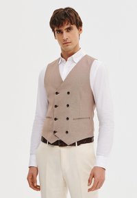 Beżowa kamizelka w wzór houndstooth z pięcioma czarnymi guzikami, slim fit, z bocznymi kieszeniami, noszona na białej koszuli oraz jasnych spodniach.