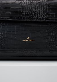Sac en cuir à texture crocodile noire avec logo doré "Anna Field" centré sur le rabat avant, posé sur une surface blanche.
