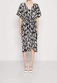 Vestido de patrones en blanco y negro con escote en V, mangas cortas y cintura con lazo. De longitud media con una abertura lateral, combinado con sandalias de plataforma blancas.