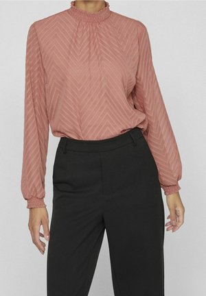 Femme portant un blouse texturé à manches longues de couleur rose poussiéreux avec un motif en chevrons, rentrée dans un pantalon noir taille haute et ajusté.