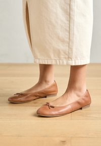 Pies con bailarinas de cuero de color marrón claro con pequeños lazos, combinados con pantalones cortos beige sobre un fondo de suelo de madera.