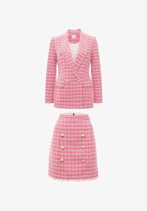 Różowy tweedowy garnitur składający się z podwójnie zapiętej marynarki z złotymi guzikami oraz koordynującej spódnicy z frędzlami i dopasowanymi akcentami guzika.