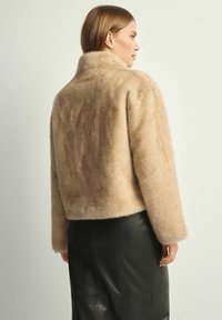 Beżowa kurtka z sztucznego futra z wysokim kołnierzem, krótki krój i gładka faktura. Nosi się ją na czarnej plisowanej spódnicy, prezentując minimalistyczny styl.