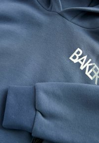 Felpa con cappuccio blu realizzata in tessuto morbido, con il logo "BAKER" stampato in bianco sul petto e polsini elasticizzati.