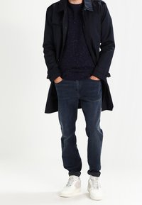 Dark navy trench coat over a navy speckled sweater, paired with blue jeans and white sneakers featuring light accents.