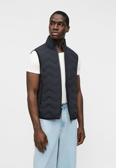 Ενήλικας άνδρας που φοράει μαύρο αμάνικο γιλέκο με ραφές πάνω από λευκό μπλουζάκι και ανοιχτόχρωμα μπλε τζιν, στέκεται μπροστά σε απλό λευκό φόντο.