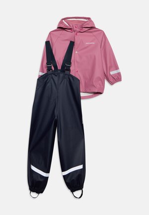 Veste à capuche rose avec bandes réfléchissantes, associée à un pantalon salopette noir imperméable avec des bretelles réglables et des guêtres aux chevilles pour enfants.