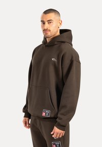 Brązowy hoodie z miękkiego materiału, z przednią kieszenią kangurką i małym logo na piersi. W komplecie z dopasowanymi spodniami.