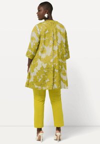 Donna in piedi di lato, che indossa una tunica floreale gialla, pantaloni gialli uniti, tacchi alti beige e grandi orecchini rotondi bianco e nero.