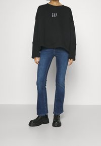 Person klädd i en svart GAP-tröja, blå bootcut jeans och svarta kängor med tjock sula, stående mot en enkel bakgrund.