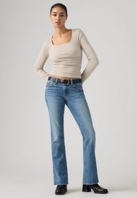 Γυναίκα που στέκεται με τα χέρια πίσω από την πλάτη, ντυμένη με μπεζ μακρυμάνικη μπλούζα, μπλε τζιν με ίσια μπατζάκια, μαύρη ζώνη και μαύρες μπότες με τακούνι.