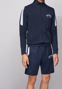 Chándal marino con chaqueta con cremallera y pantalones cortos, que presenta rayas laterales en blanco y el logo "BOSS", fabricado en un tejido suave y texturizado.