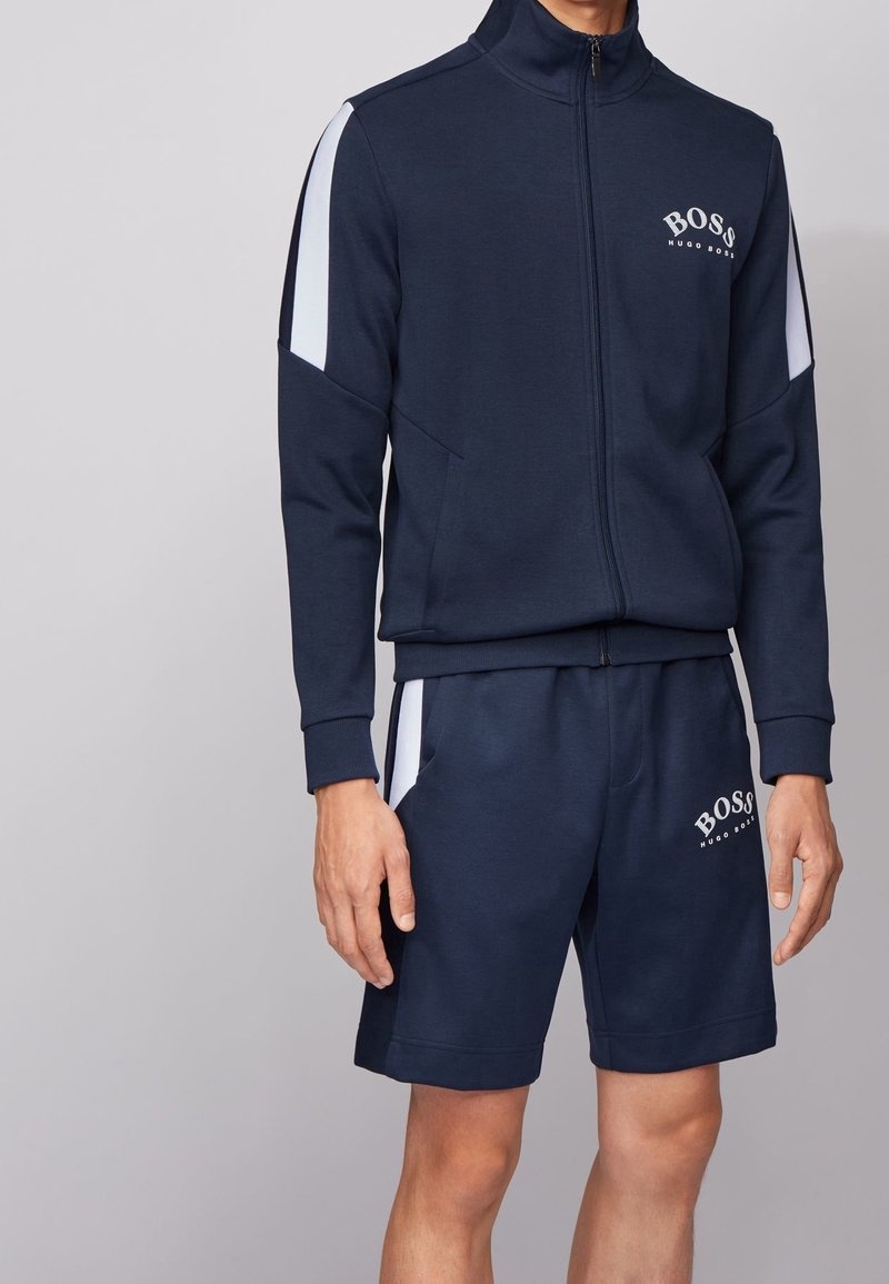 Chándal marino con chaqueta con cremallera y pantalones cortos, que presenta rayas laterales en blanco y el logo "BOSS", fabricado en un tejido suave y texturizado.