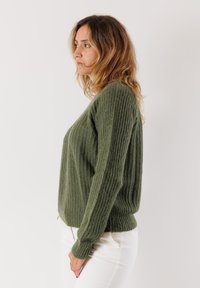 Donna con capelli ondulati che indossa un maglione verde lavorato a maglia e pantaloni bianchi, in piedi di profilo contro uno sfondo chiaro e semplice.