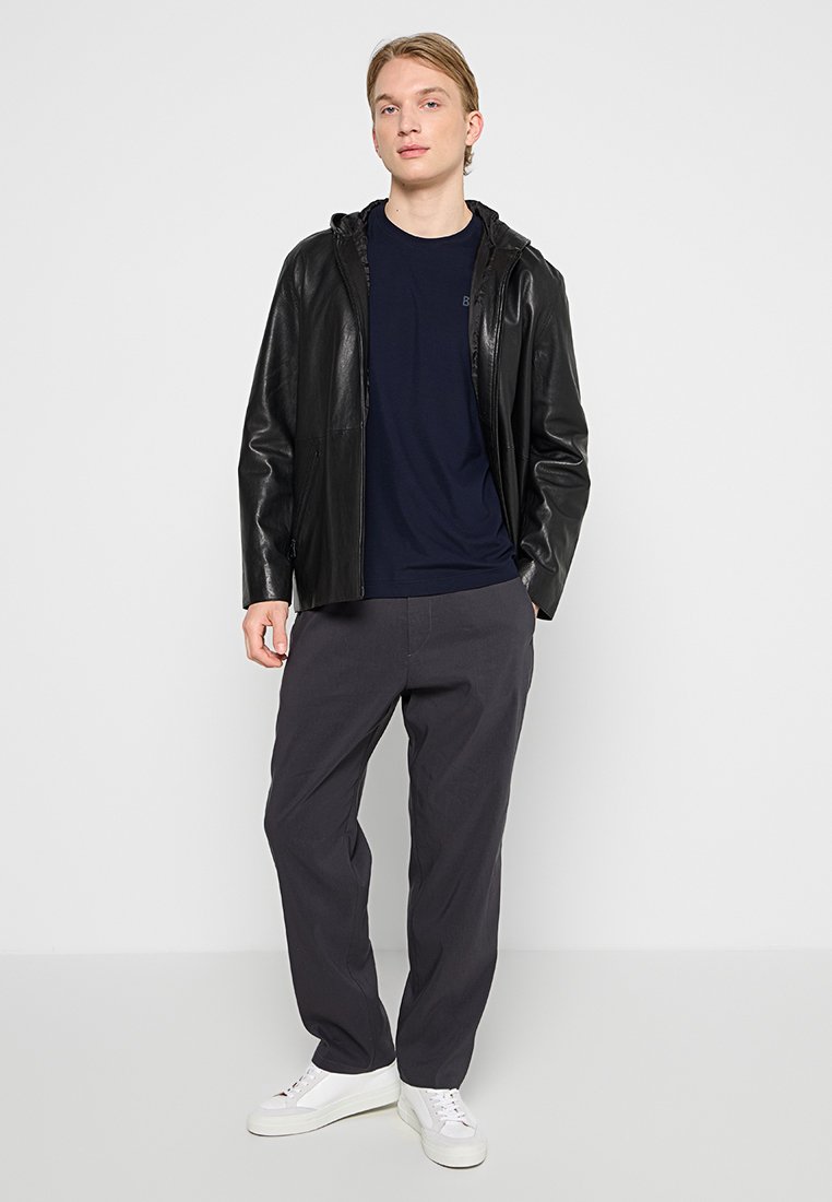 Schwarze Lederjacke mit Kapuze über einem dunkelblauen T-Shirt, kombiniert mit lockeren grauen Hosen und weißen Sneakers. Einfaches Design, strukturierter Stoff.