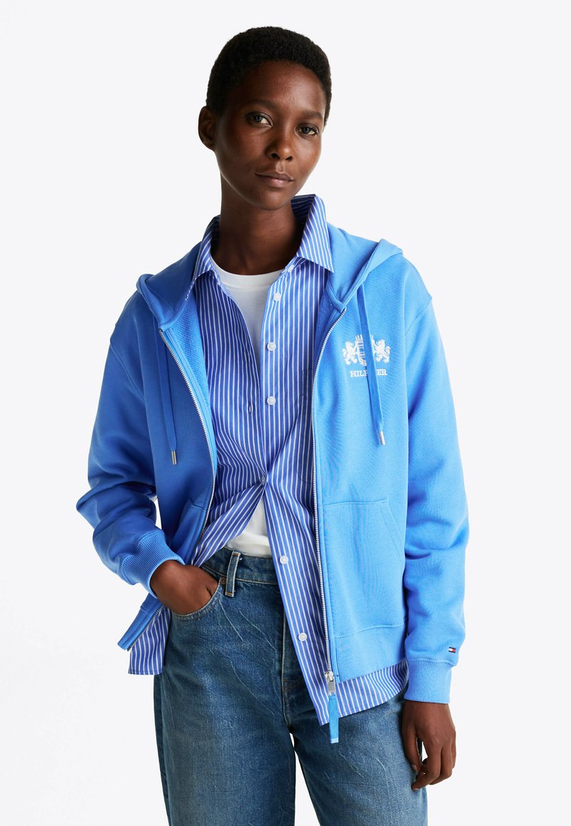Sudadera azul de algodón con cremallera y bolsillo tipo canguro, combinada con una camisa de botones a rayas azul y blanca, y pantalones vaqueros.