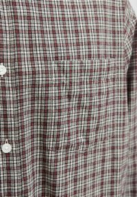 Gros plan sur le devant d'une chemise avec un petit motif à carreaux noir, blanc et rouge, poche poitrine visible et boutons blancs.